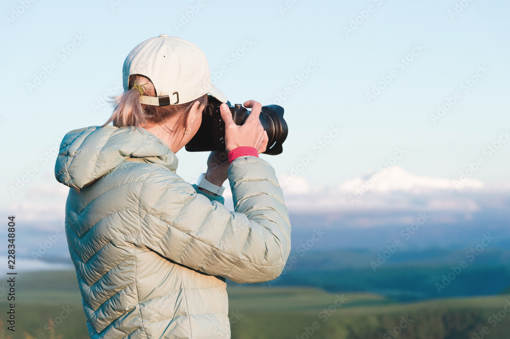 Obraz premium Portrait of a girl photographer in a cap on nature photographing on her digital mirror camera. Back view