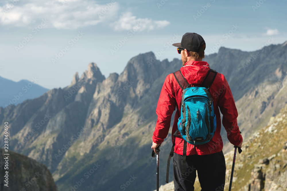 A bearded man in sunglasses and a cap with a backpack stands on top of a rock and looks into a rocky valley high in the mountains. The concept of tourism and easy trekking in the mountains outdoor