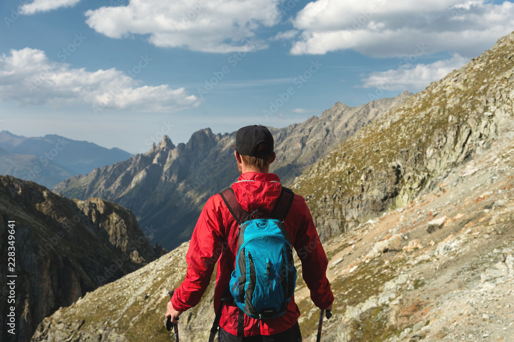 A bearded man in sunglasses and a cap with a backpack stands on top of a rock and looks into a rocky valley high in the mountains. The concept of tourism and easy trekking in the mountains outdoor