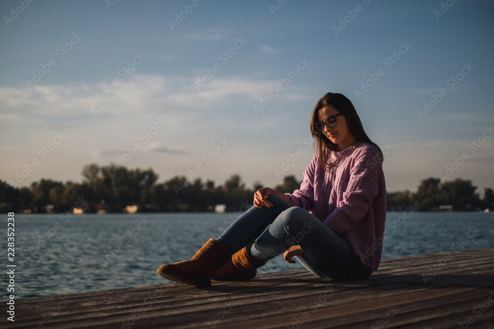 Lonely girl with glasses sitting by the river in sunset