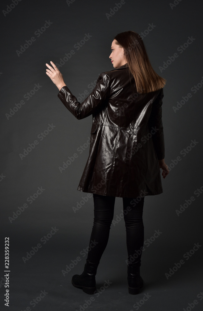 full length portrait of brunette woman wearing long leather coat. standing pose with back to the camera, on grey studio background. full length portrait of brunette woman wearing long leather coat. standing pose with back to the camera, on grey studio background.