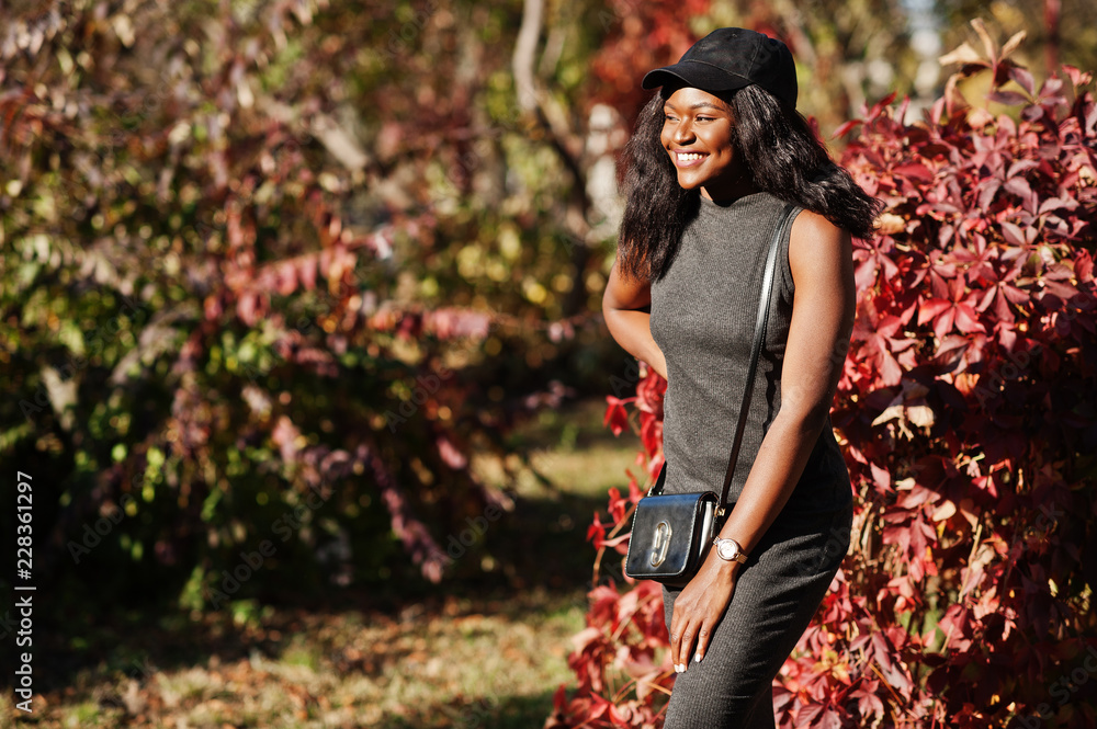 Stylish african american girl in gray tunic, crossbody bag and cap posed at sunny autumn day against red leaves. Africa model woman. Stylish african american girl in gray tunic, crossbody bag and cap posed at sunny autumn day against red leaves. Africa model woman.