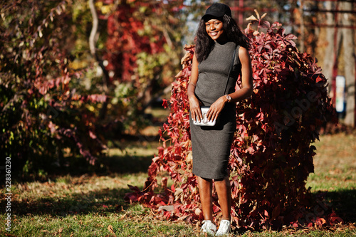 Stylish african american girl in gray tunic, crossbody bag and cap posed at sunny autumn day against red leaves. Africa model woman. photo