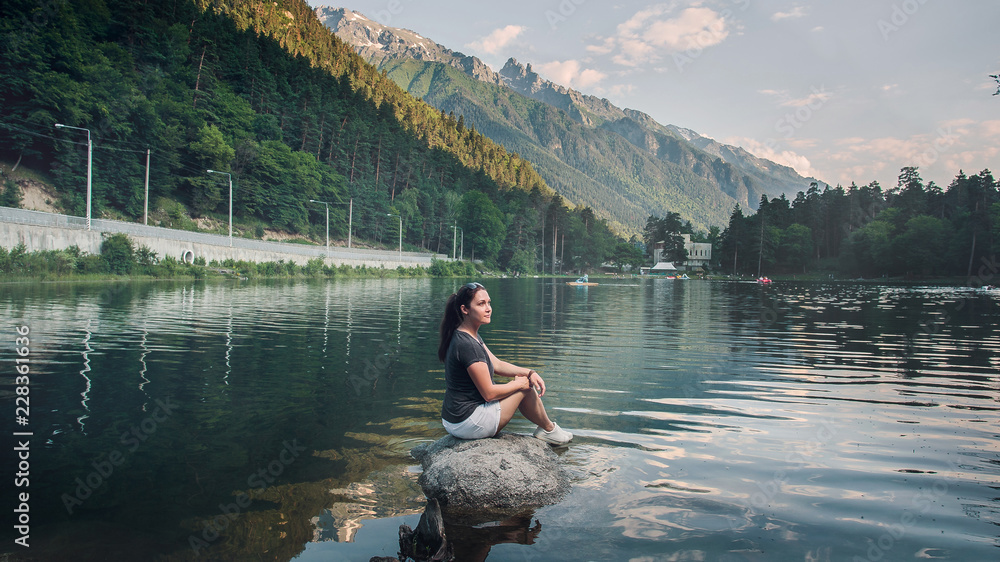 European girl tourist sitting on a stone in the middle of the pond on the background of high mountains covered with green forest. Summer, evening. Caucasus, Russia.