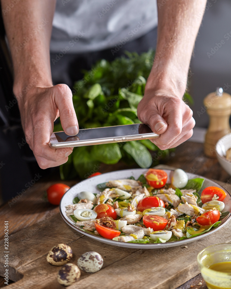 Appetizing healthy salad from organic ingredients in a plate with mens hand making the photo by smartphon on a wooden table. Appetizing healthy salad from organic ingredients in a plate with mens hand making the photo by smartphon on a wooden table.