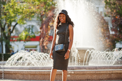 Stylish african american girl in gray tunic, crossbody bag and cap posed at sunny autumn day against fountains. Africa model woman. photo