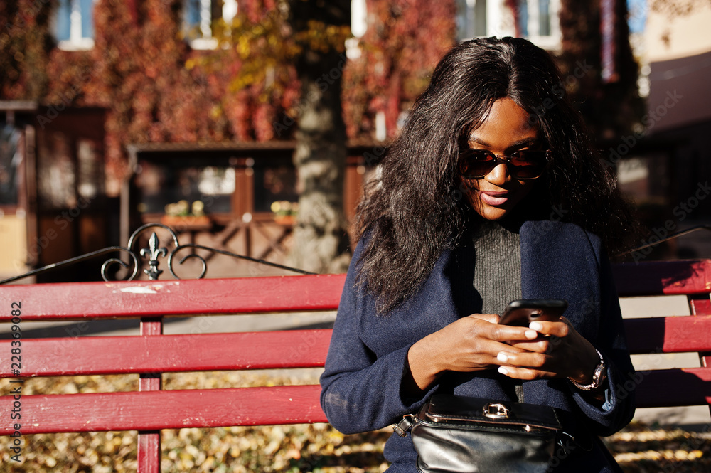 Fototapeta premium Stylish african american girl in blue coat and sunglasses posed at sunny autumn day at bench and using her mobile phone. Africa model woman with smartphone.