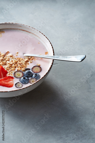 White bowl with homemade granola, sliced strawberries, yogurt, blueberry, spoon - natural breakfast on a gray table.