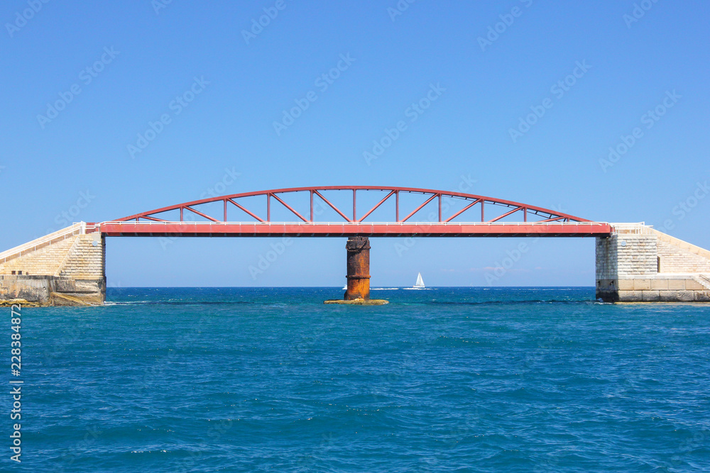 Small white sailboat ship in blue water of Grand Harbor, Valletta. Red Breakwater Bridge metallic construction on foreground