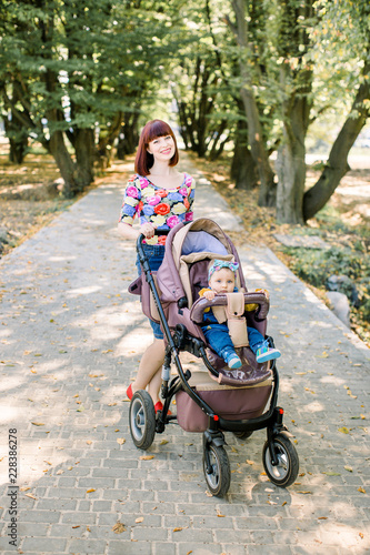 Young mother walking with smiling baby in brown stroller. Parents walking outdoors with child in summer pram.