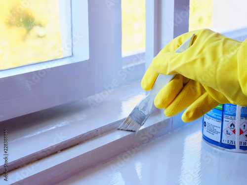 Handyman paints a window frame with white paint with a paint brush. Hand in yellow rubber gloves applies a glossy paint finish to window frame. photo