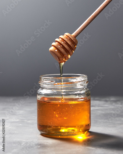 Fragrant organic fresh honey dripping from wooden stick to a glass pot on a gray marble table, pure natural sweet goodness.