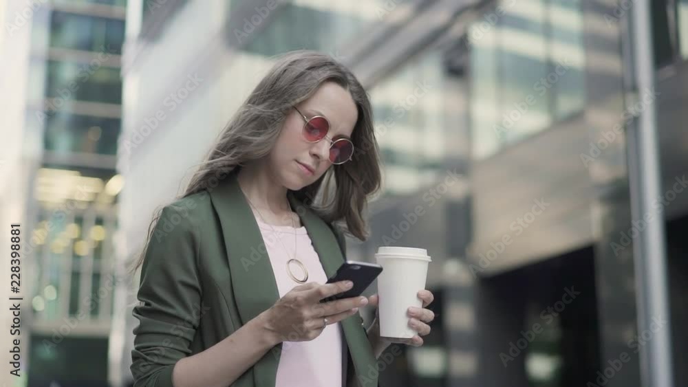 Young girl in khaki jacket is standing in city street, web surfing and drinking coffee to go. Blurred background. Slider slow motion medium shot Young girl in khaki jacket is standing in city street, web surfing and drinking coffee to go. Blurred background. Slider slow motion medium shot
