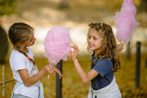 Two 7 year old girls eating cotton candy in park. Shot taken in autumn, lots of fallen leaves on ground. Cotton candy is big and pink. Twin sisters are blond, curly hair, and braids. White, navy blue.