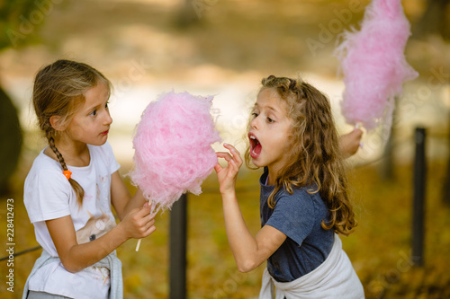 Two 7 year old girls eating cotton candy in park. Shot taken in autumn, lots of fallen leaves on ground. Cotton candy is big and pink. Twin sisters are blond, curly hair, and braids. White, navy blue.