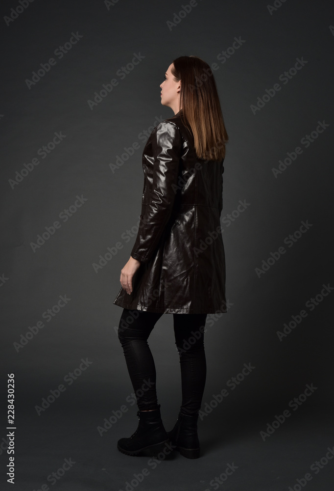 full length portrait of brunette woman wearing long leather coat. standing pose with back to the camera, on grey studio background. full length portrait of brunette woman wearing long leather coat. standing pose with back to the camera, on grey studio background.