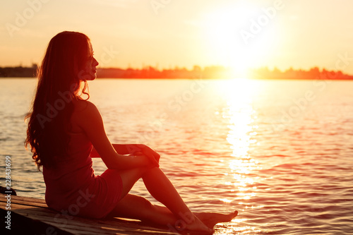 Romantic girl sitting on the pier near the water at sunset. Woman by the sea. Model on the beach on a background of sunlight. Siluet in backlight.