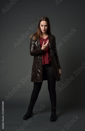 full length portrait of brunette girl wearing long leather coat and boots. standing pose on grey studio background.