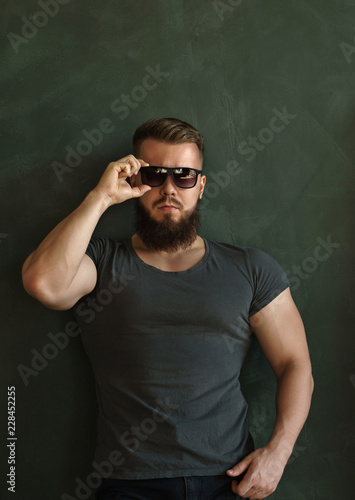 Bodybuilder in sunglasses portrait. Muscular man with a beard in a tight t-shirt straightens his glasses. Stylish and sporty.
