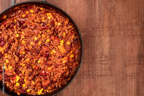 A closeup photo of a large cast iron pan with chili con carne, shot from above on a dark rustic wooden background with a place for text
