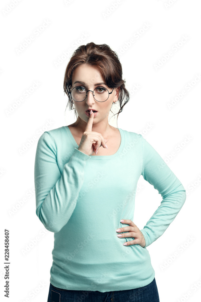Slightly puzzled expression. A young brown-haired woman in a mint-colored jumper and in round glasses narrowed her eyes and held her index finger at the lips, calling for silence. Slightly puzzled expression. A young brown-haired woman in a mint-colored jumper and in round glasses narrowed her eyes and held her index finger at the lips, calling for silence.