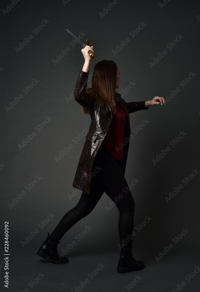 full length portrait of brunette girl wearing long leather coat and boots. standing pose and holding a sword on grey studio background. full length portrait of brunette girl wearing long leather coat and boots. standing pose and holding a sword on grey studio background.