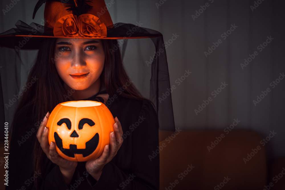 Attractive beautiful Asian woman dressed as a witch holding jack-o-lantern on hand, Halloween holidays in cinematic dark tone. 