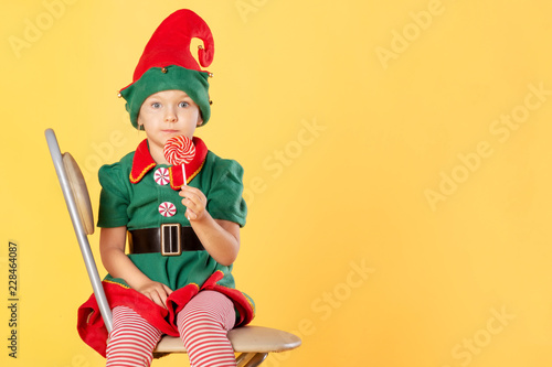 A beautiful baby in a suit of a Christmas elf is sitting on a chair. She has a delicious lollipop in her hands. Surprise. Near Copy Space.