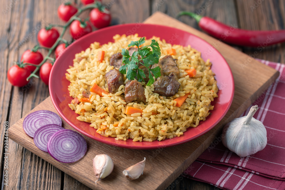 Red plate with pilaf on a brown wooden table. On the table is red pepper, green onions, garlic, cherry tomatoes, red napkin, spoon. Red plate with pilaf on a brown wooden table. On the table is red pepper, green onions, garlic, cherry tomatoes, red napkin, spoon.