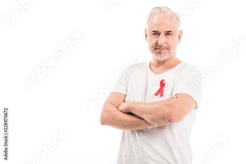 handsome mature man in blank white t-shirt with aids awareness red ribbon looking at camera with crossed arms isolated on white