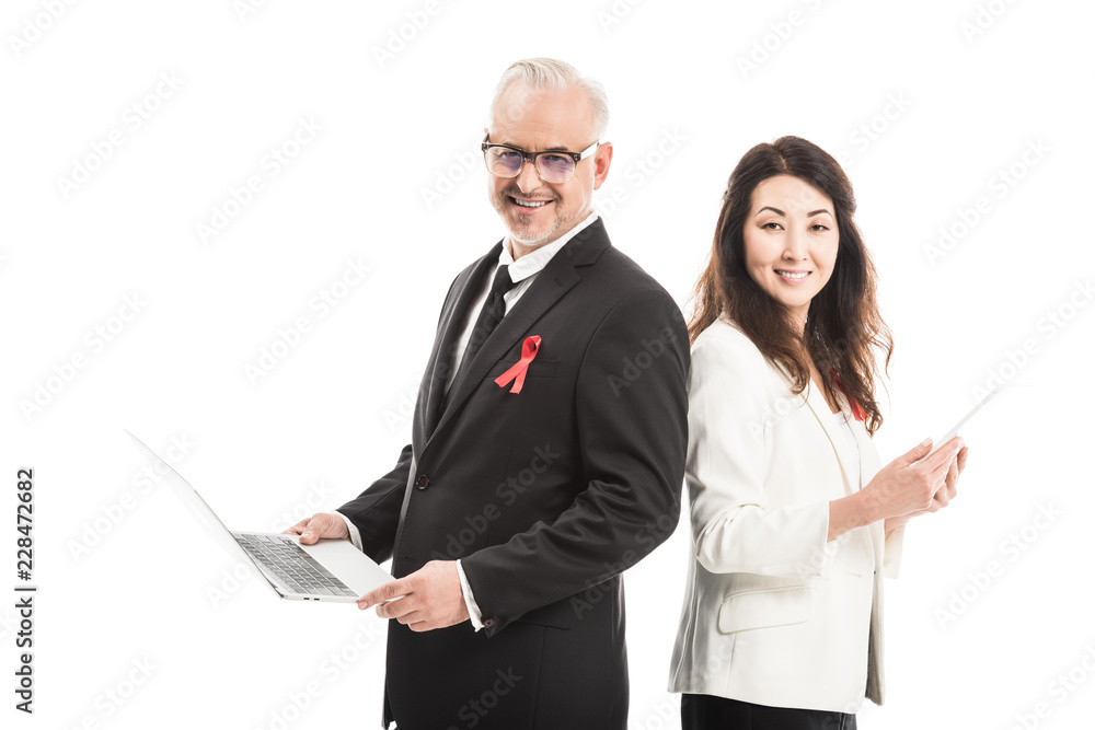 Fototapeta premium multiethnic happy businesspeople with aids awareness red ribbons working with gadgets while standing back to back and looking at camera isolated on white