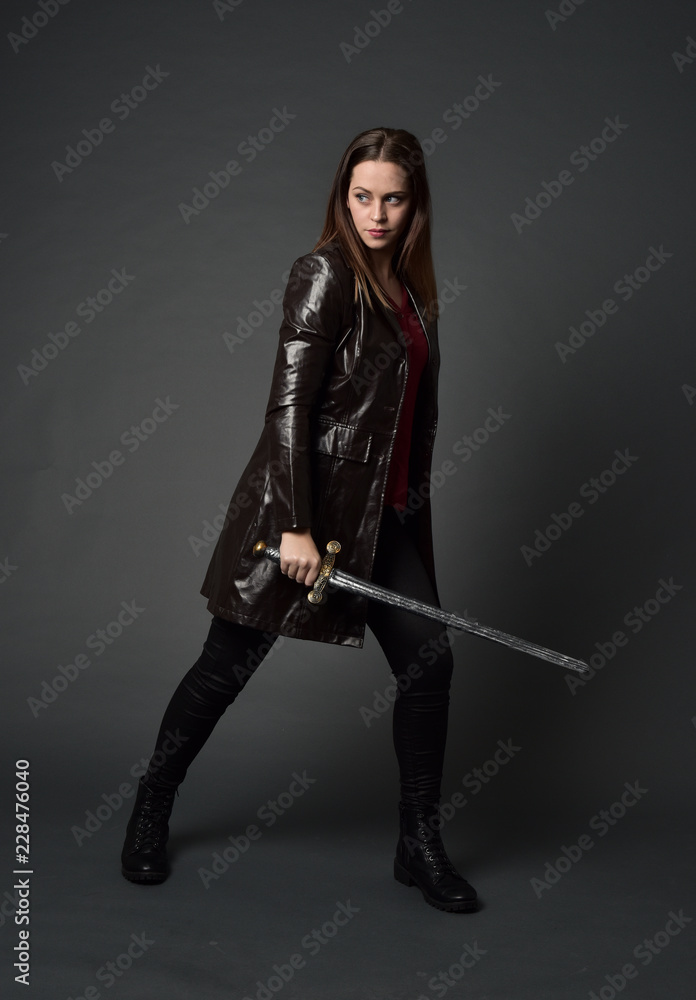 full length portrait of brunette girl wearing long leather coat and boots. standing pose and holding a sword on grey studio background. full length portrait of brunette girl wearing long leather coat and boots. standing pose and holding a sword on grey studio background.