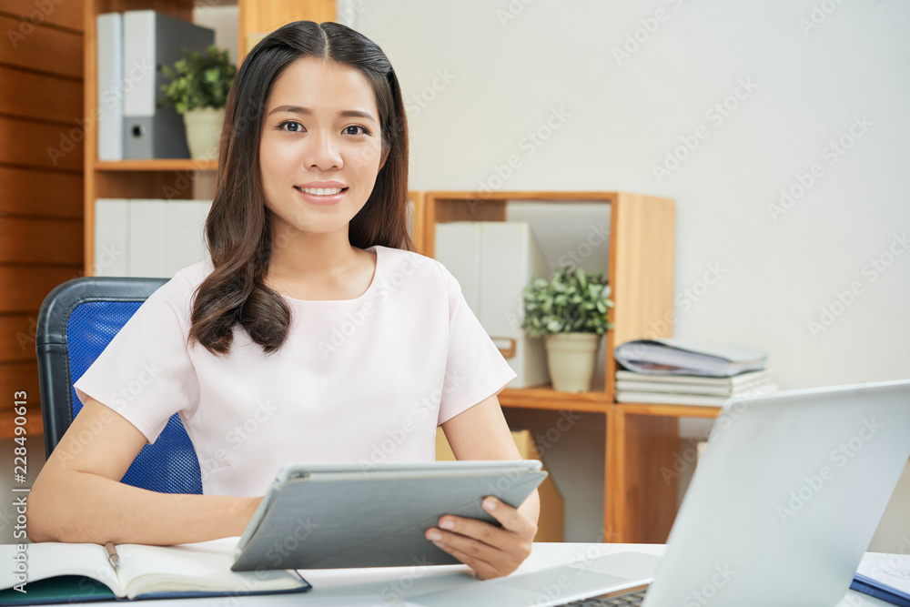 Beautiful young Asian woman working in office sitting at table with tablet and smiling at camera