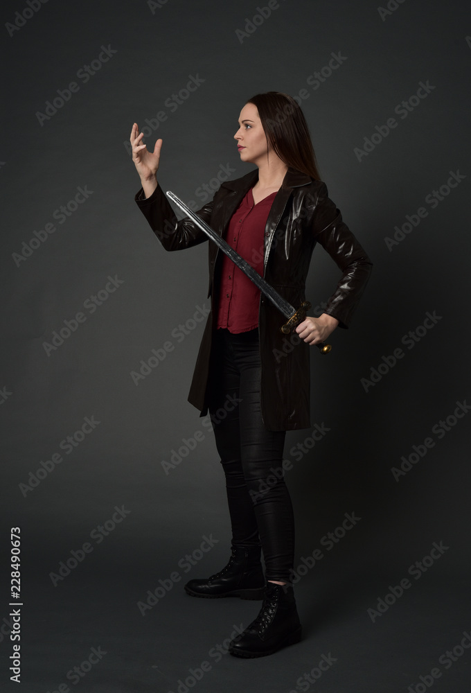 full length portrait of brunette girl wearing long leather coat and boots. standing pose and holding a sword on grey studio background. full length portrait of brunette girl wearing long leather coat and boots. standing pose and holding a sword on grey studio background.
