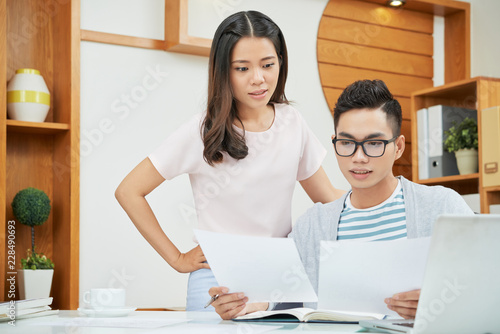 Modern Asian woman and man gathering at table in office and looking at paper documents
