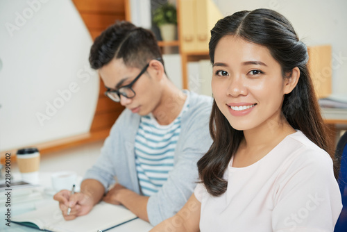 Pretty modern Asian woman at workplace with young male coworker near smiling at camera