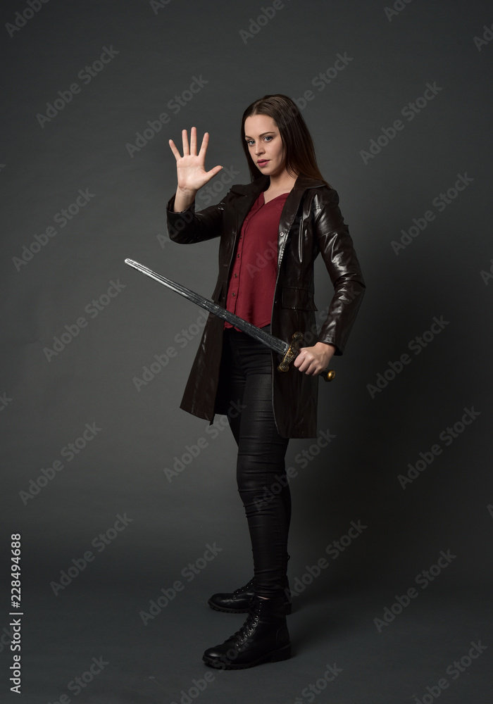 full length portrait of brunette girl wearing long leather coat and boots. standing pose and holding a sword on grey studio background. full length portrait of brunette girl wearing long leather coat and boots. standing pose and holding a sword on grey studio background.
