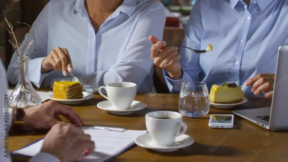 Close up shot of three unrecognizable businesspeople sitting at table in cafe or restaurant, eating cakes and drinking water and coffee and discussing text of agreement Close up shot of three unrecognizable businesspeople sitting at table in cafe or restaurant, eating cakes and drinking water and coffee and discussing text of agreement