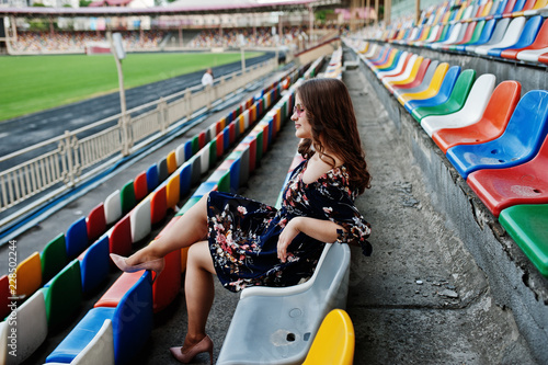 Portrait of a young beutiful girl in dress and sunglasses sitting on the tribunes in stadium.
