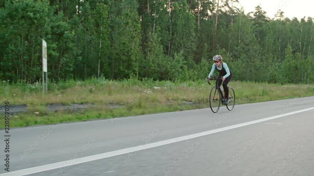 Tracking of female cyclist in protective helmet and professional sportswear riding bicycle along road surrounded by green forest