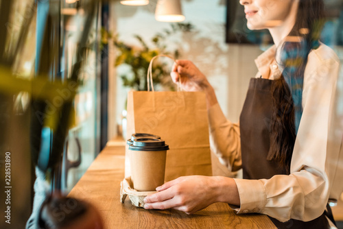 cropped image of waitress in apron holding disposable coffee cups and paper bag in cafe