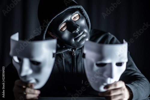 Mystery hoody man wearing black mask holding two white masks in his hand. Anonymous social masking. Major depressive disorder or bipolar disorder. Halloween concept