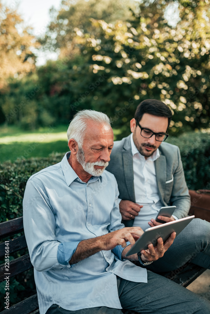 Senior businessman giving advice to a younger colleague outdoors. Looking at tablet while sitting on the bench. Senior businessman giving advice to a younger colleague outdoors. Looking at tablet while sitting on the bench.