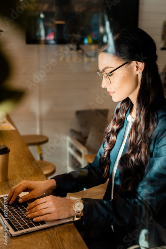 beautiful brunette teleworker in eyeglasses using laptop at restaurant