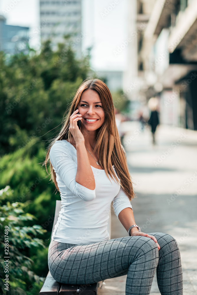 Smiling young woman talking over mobile phone while sitting on the bench. Smiling young woman talking over mobile phone while sitting on the bench.