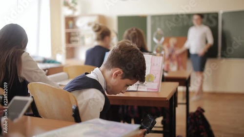 A bad student is playing with smartphone during class under the Desk, hiding from the teacher. The teacher observes how the student is note-taking lecture plays on a smartphone.