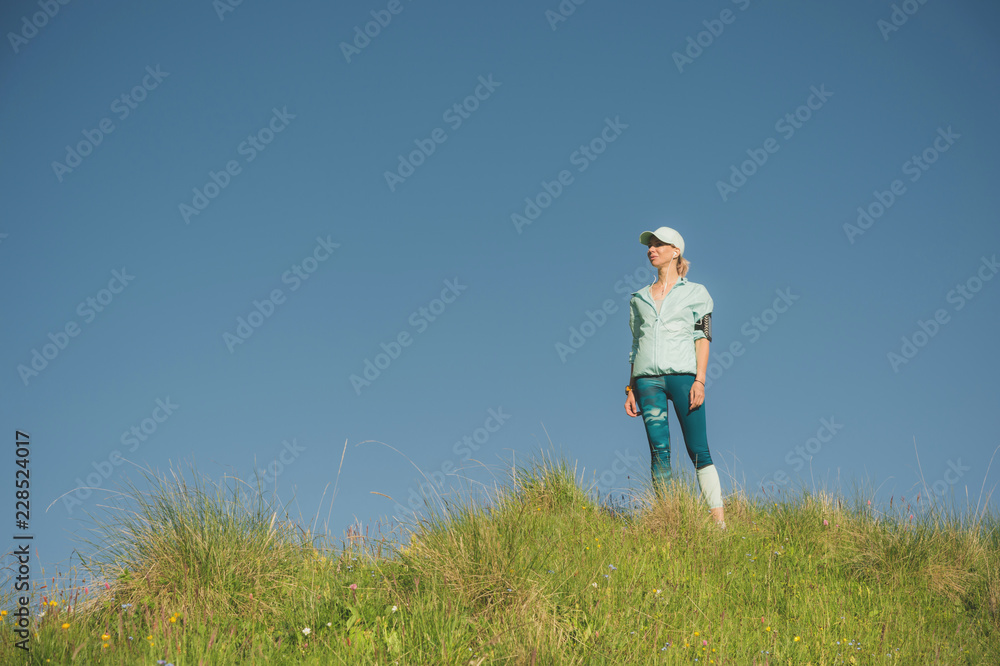 Fitness runner woman listening to music on the nature. Portrait of beautiful girl wearing earphones earbuds and running cap. against the background of a green field and blue sky