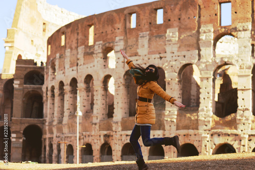 Young woman in winter clothes having fun on the background of the Winter Colosseum in Rome. The girl happily bounces on the photo of the Colosseum. Celebration of the New Year in Rome