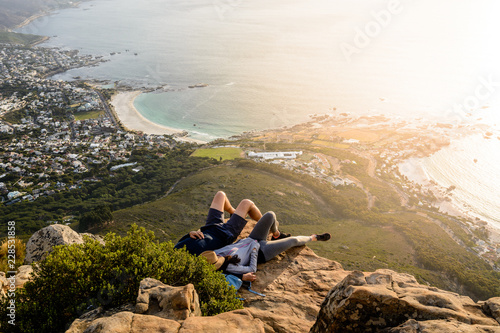 A couple is relaxing on top of Lion's Head mountain in Cape Town and enjoying the beautiful sunset with a view of Camps Bay and Clifton Beach areas
