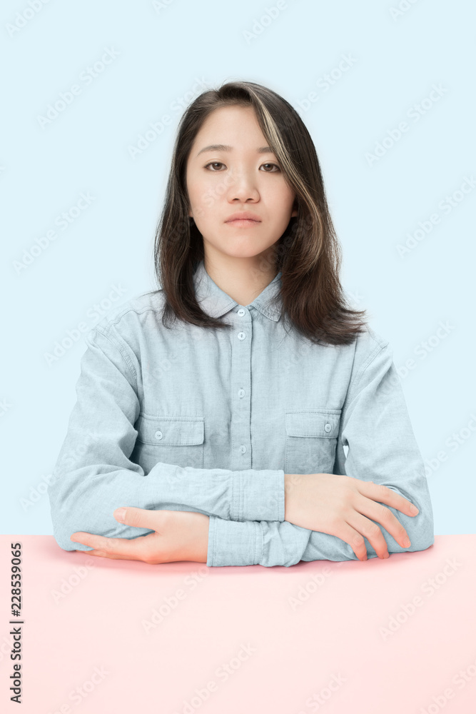Serious business woman sitting at table, looking at camera isolated on trendy blue studio background. Beautiful, young face. Female half-length portrait. Human emotions, facial expression concept. Serious business woman sitting at table, looking at camera isolated on trendy blue studio background. Beautiful, young face. Female half-length portrait. Human emotions, facial expression concept.